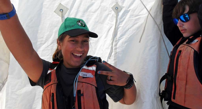 A person wearing a life jacket stands in front of a sail and gives the camera a peace sign